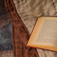 Hand-stitched linen quilt in oatmeal and cinnamon brown draped over a sofa with an open book, showing the hand-stitched cross and dash pattern up close