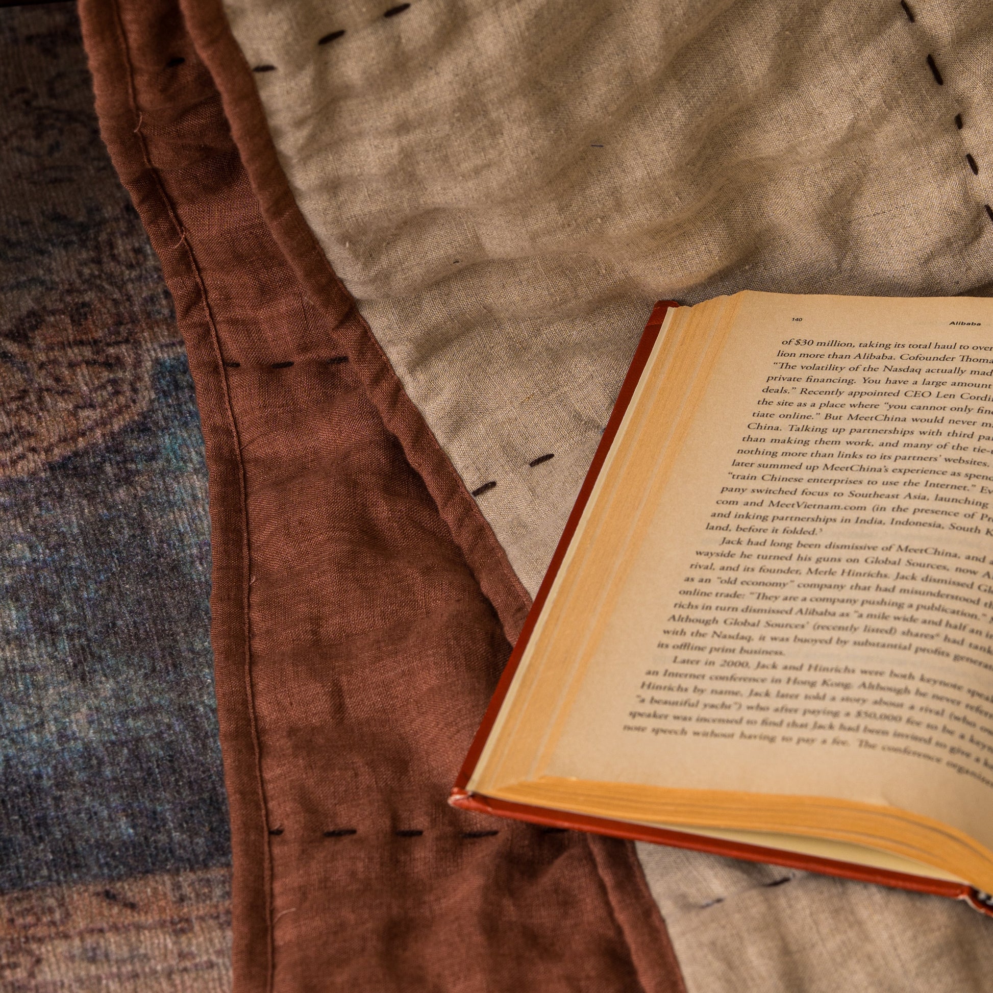 Hand-stitched linen quilt in oatmeal and cinnamon brown draped over a sofa with an open book, showing the hand-stitched cross and dash pattern up close