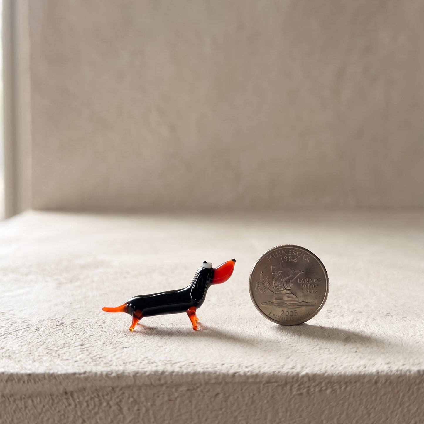The Little Sausage miniature glass dachshund shown next to a U.S. quarter for scale, lampworked in solid Ukrainian glass