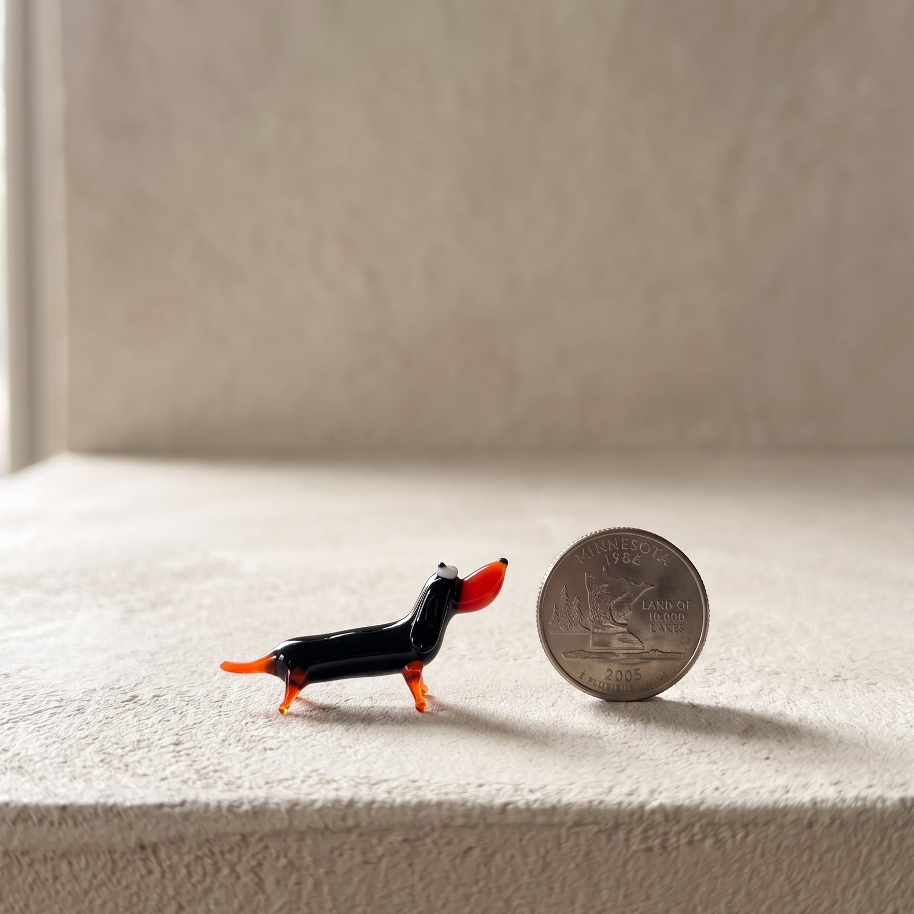 The Little Sausage miniature glass dachshund shown next to a U.S. quarter for scale, lampworked in solid Ukrainian glass