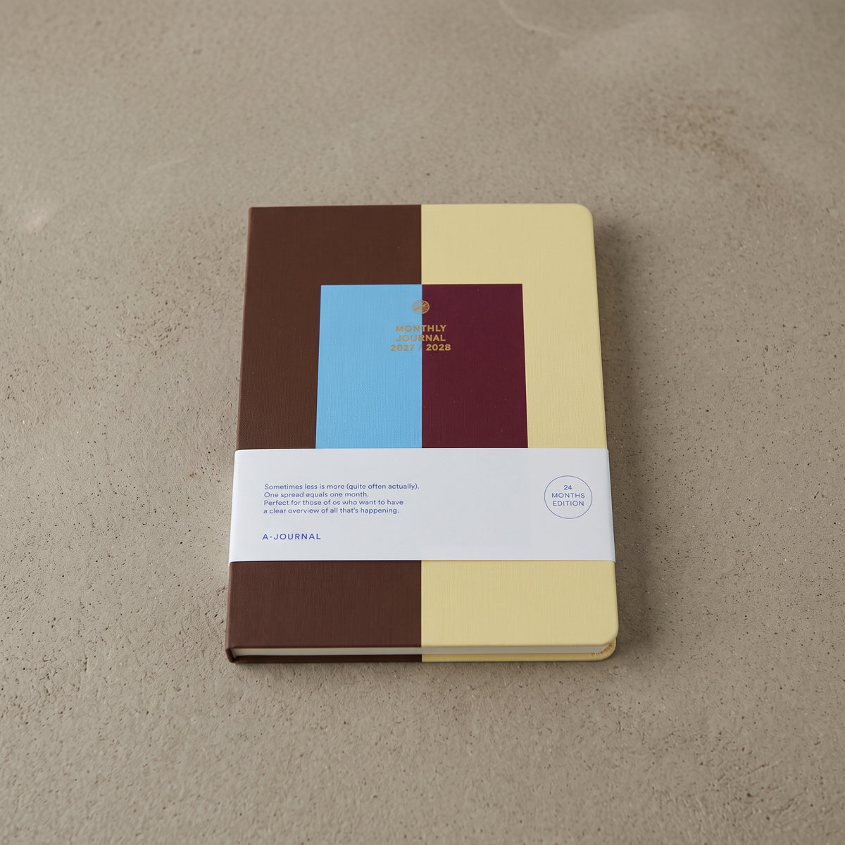 Two-year monthly journal with a color-block cover in chocolate brown, butter yellow, sky blue and burgundy, gold-foil lettering reading Monthly Journal 2027 / 2028, shown with its paper belly band