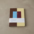 Two-year monthly journal with a color-block cover in chocolate brown, butter yellow, sky blue and burgundy, gold-foil lettering reading Monthly Journal 2027 / 2028, shown with its paper belly band