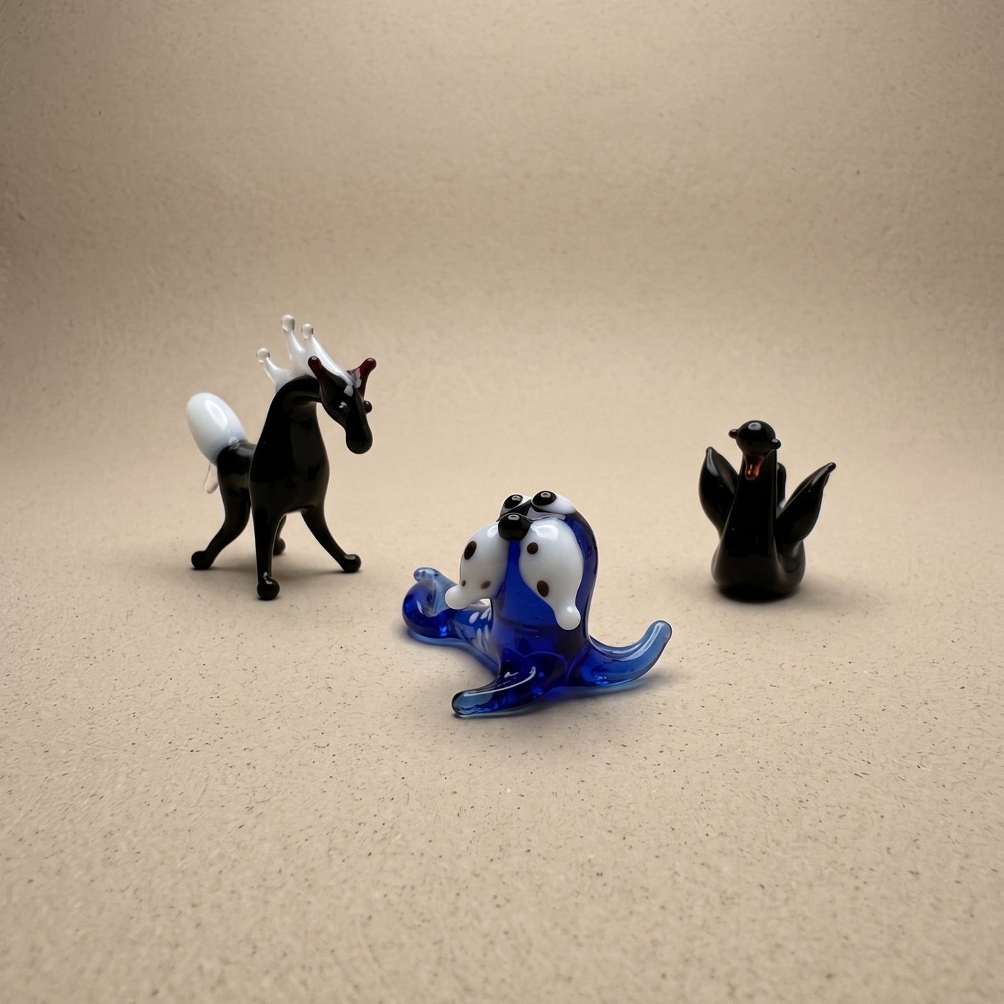 Three miniature lampworked glass figurines — black horse, blue spotted seal, and dark seal — showing variety of form and glasswork detail, by Glass by Iness