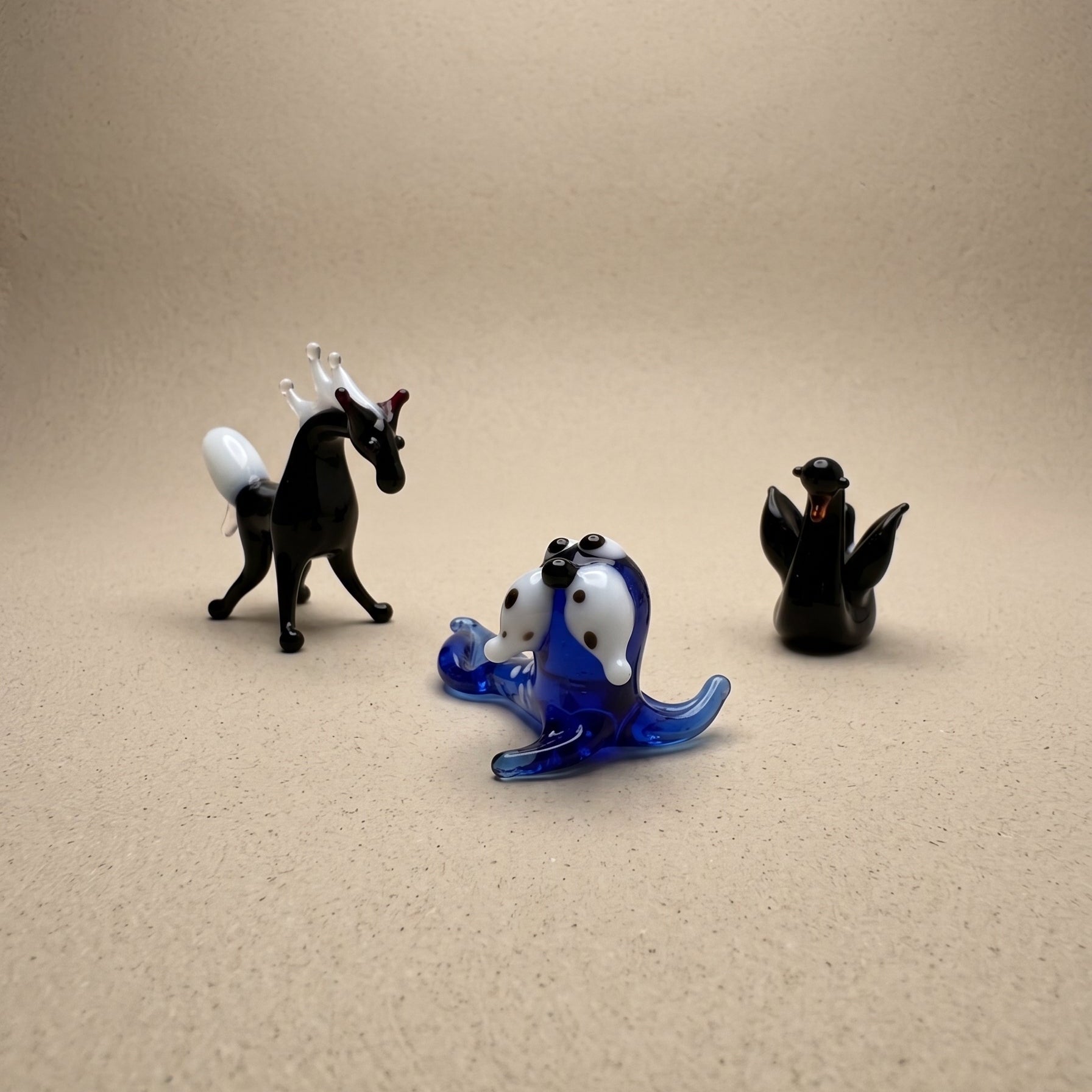 Three miniature lampworked glass figurines — black horse, blue spotted seal, and dark seal — showing variety of form and glasswork detail, by Glass by Iness
