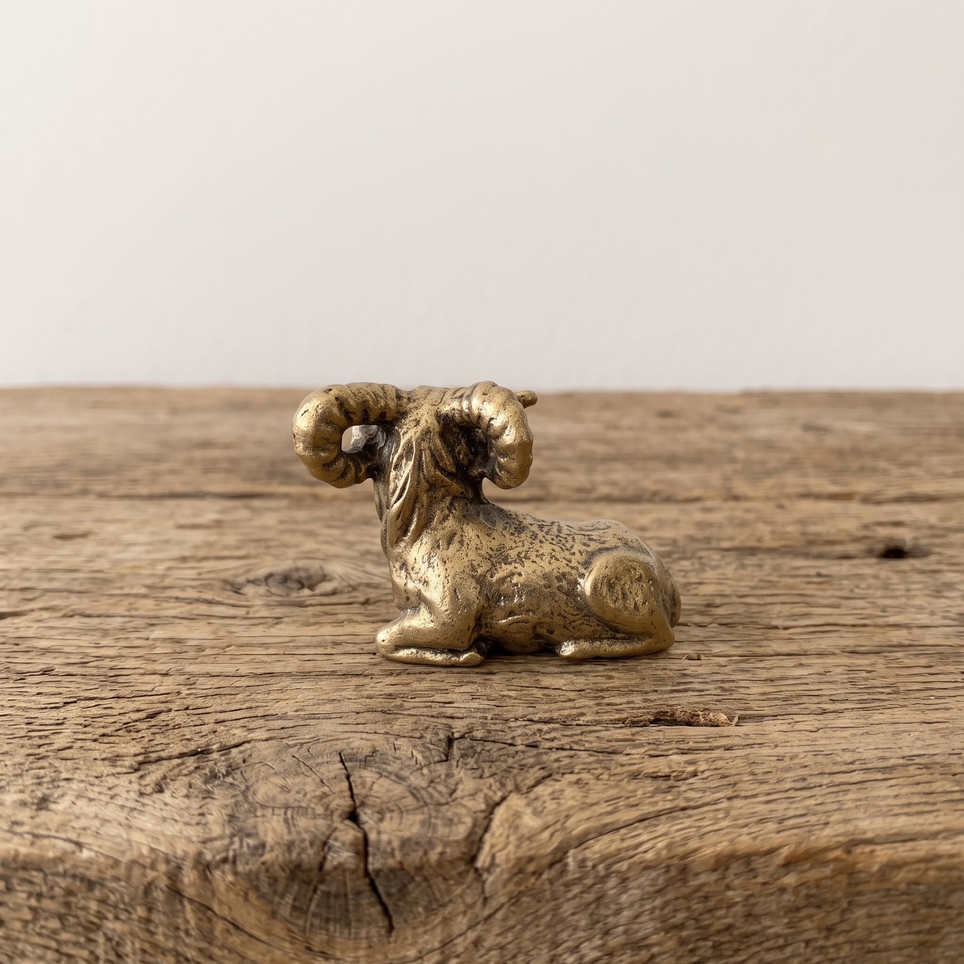Reclining brass ram figurine with curled horns and antique patina, displayed on a rustic wooden surface