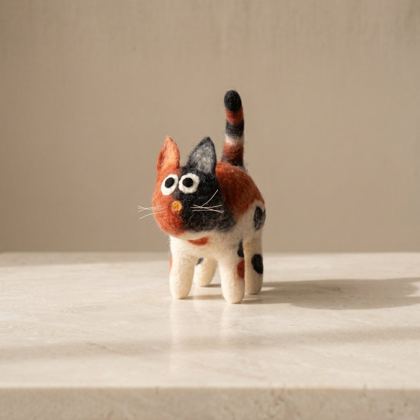 Hand-felted calico cat soft toy with ginger, black and cream wool patches, big eyes and a striped tail, made in Nepal