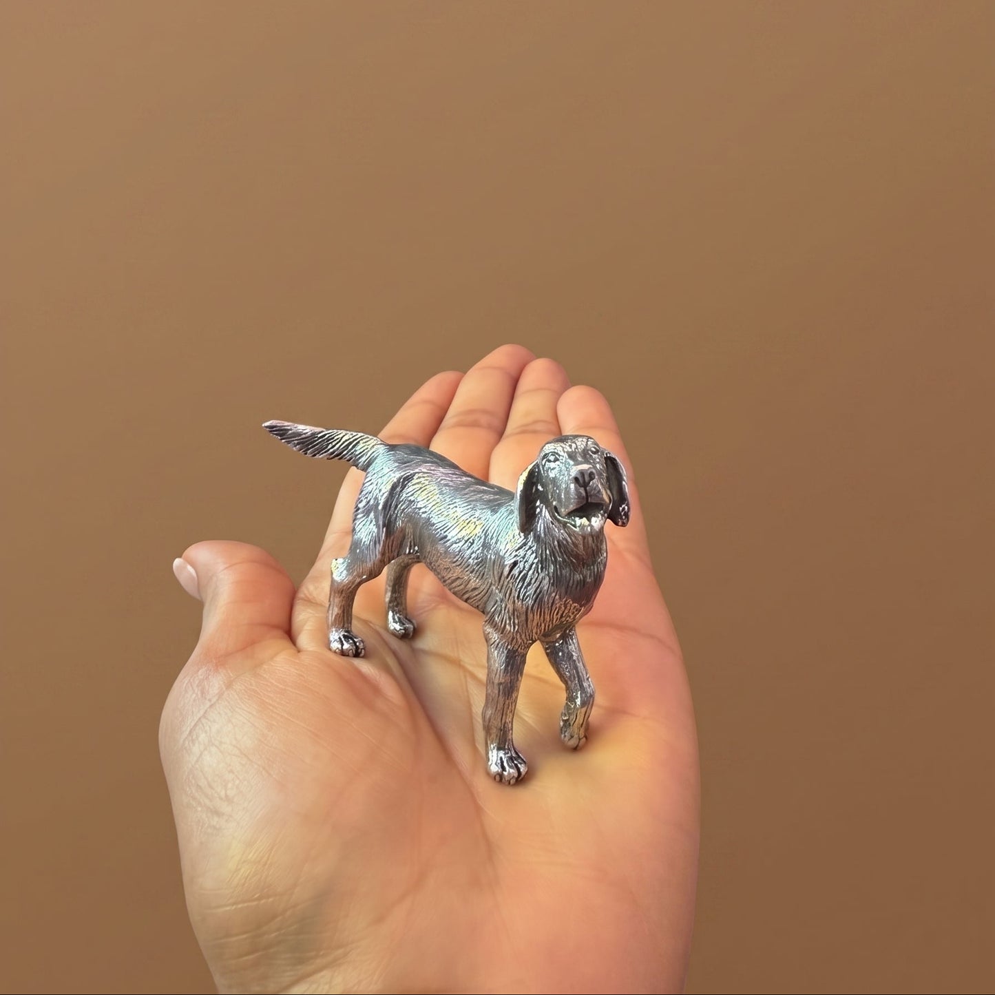 Pewter retriever figurine standing in an open palm, tail raised, showing its compact size and silver luster