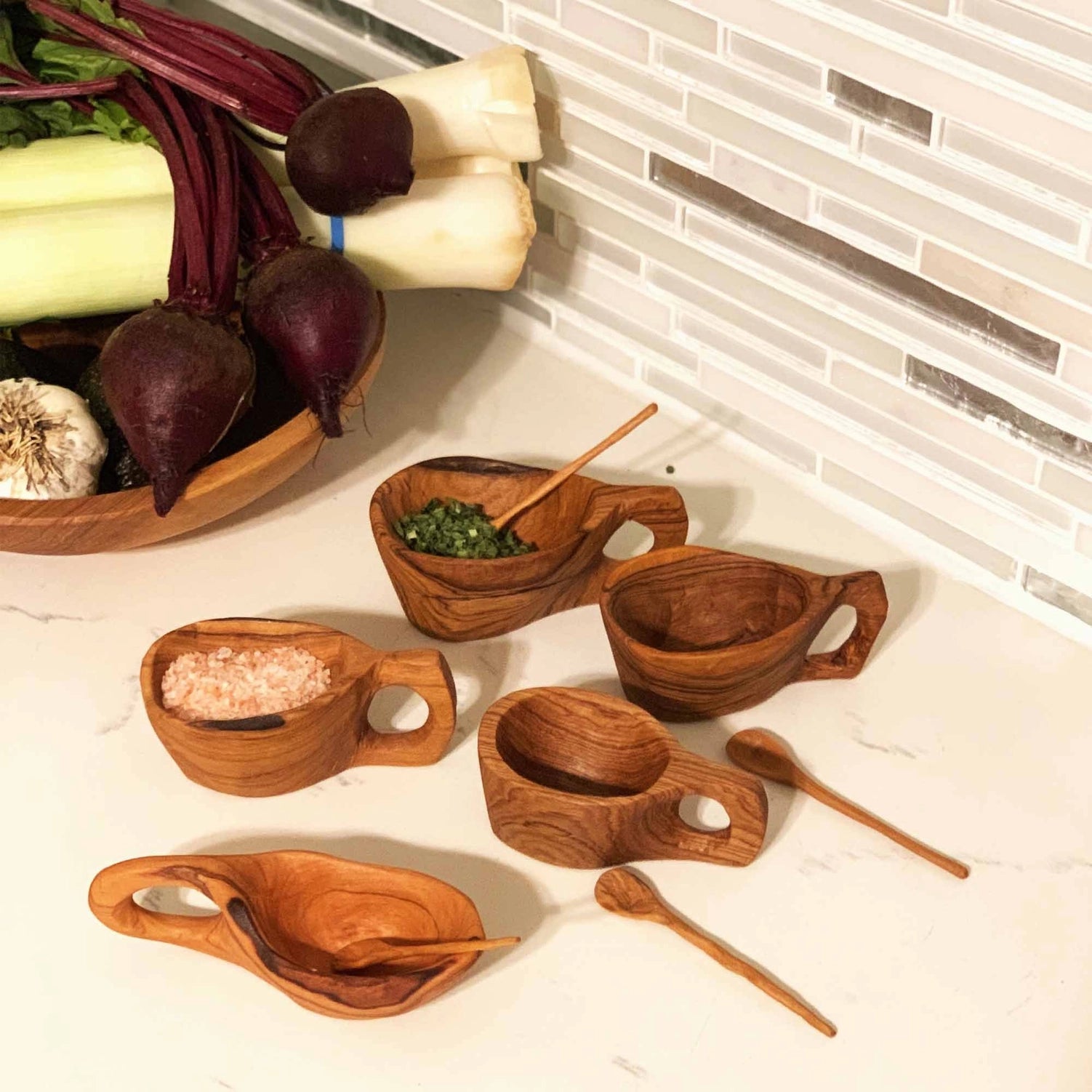 Collection of reclaimed olive wood salt pots with handles and spoons on marble kitchen counter showing unique grain variations