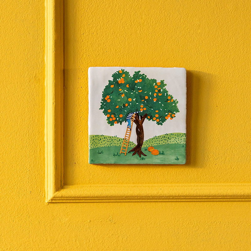 Orange Harvest Storytile displayed on a yellow wall showing illustrated orange tree with woven baskets, hand-baked ceramic by Marga van Oers in the Netherlands, from WILLAGE