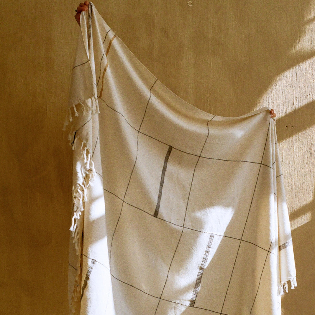 Hand-loomed organic cotton throw hanging in natural light, showing weave texture and hand-knotted fringe, from Leila Atelier