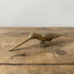 The Patient Prober miniature brass long billed bird with elongated beak and sturdy stance, showing warm aged patina on a wooden surface, from the WILLAGE Tiny Objects collection