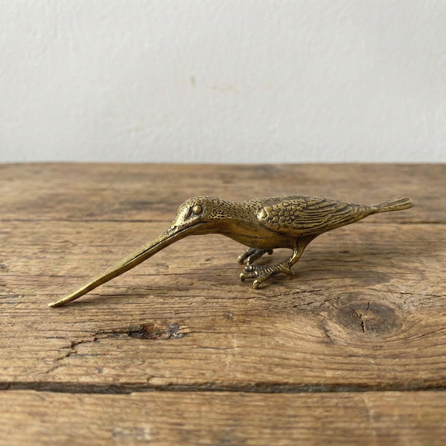 The Patient Prober miniature brass long billed bird with elongated beak and sturdy stance, showing warm aged patina on a wooden surface, from the WILLAGE Tiny Objects collection