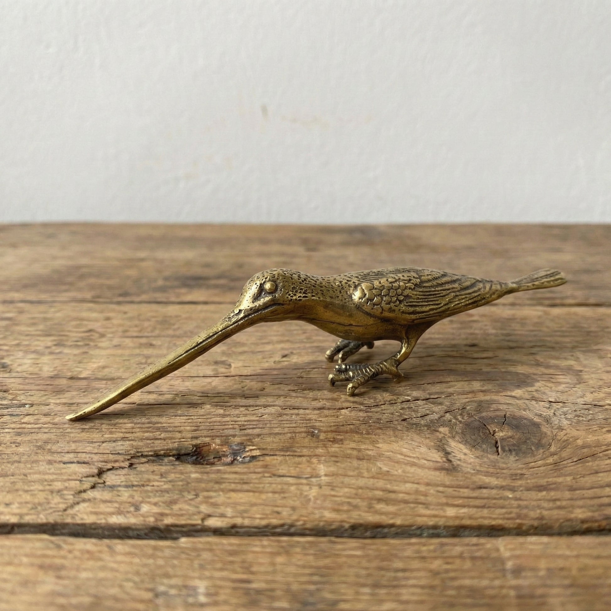 The Patient Prober miniature brass long billed bird with elongated beak and sturdy stance, showing warm aged patina on a wooden surface, from the WILLAGE Tiny Objects collection