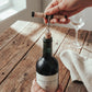 The Rattan Corkscrew being used to pull a cork from a wine bottle on a rustic wood table, showing its compact pull-style size