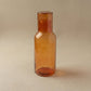 Hand-hammered recycled glass carafe in amber with tumbler lid, showing warm golden tones and hammered facets