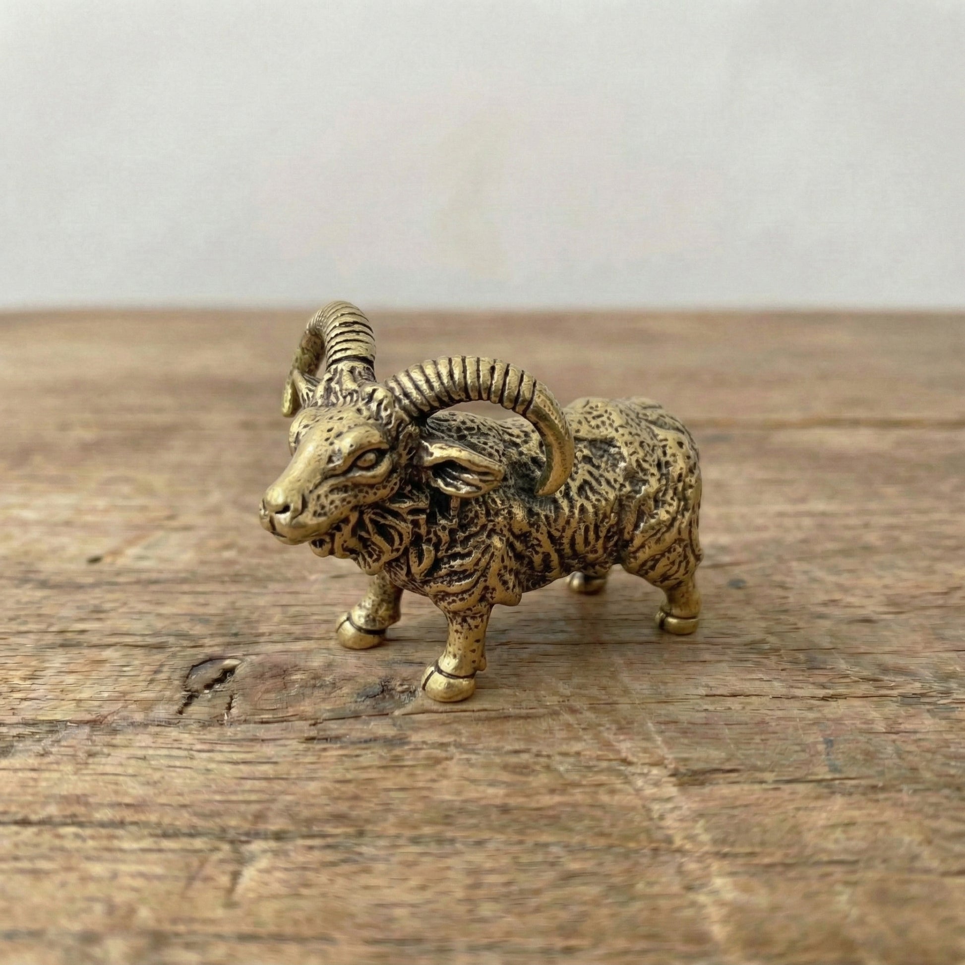 The Resilient Presence miniature brass ram with coiled horns standing firm, showing warm aged patina and textured detail on a wooden surface, from the WILLAGE Tiny Objects collection