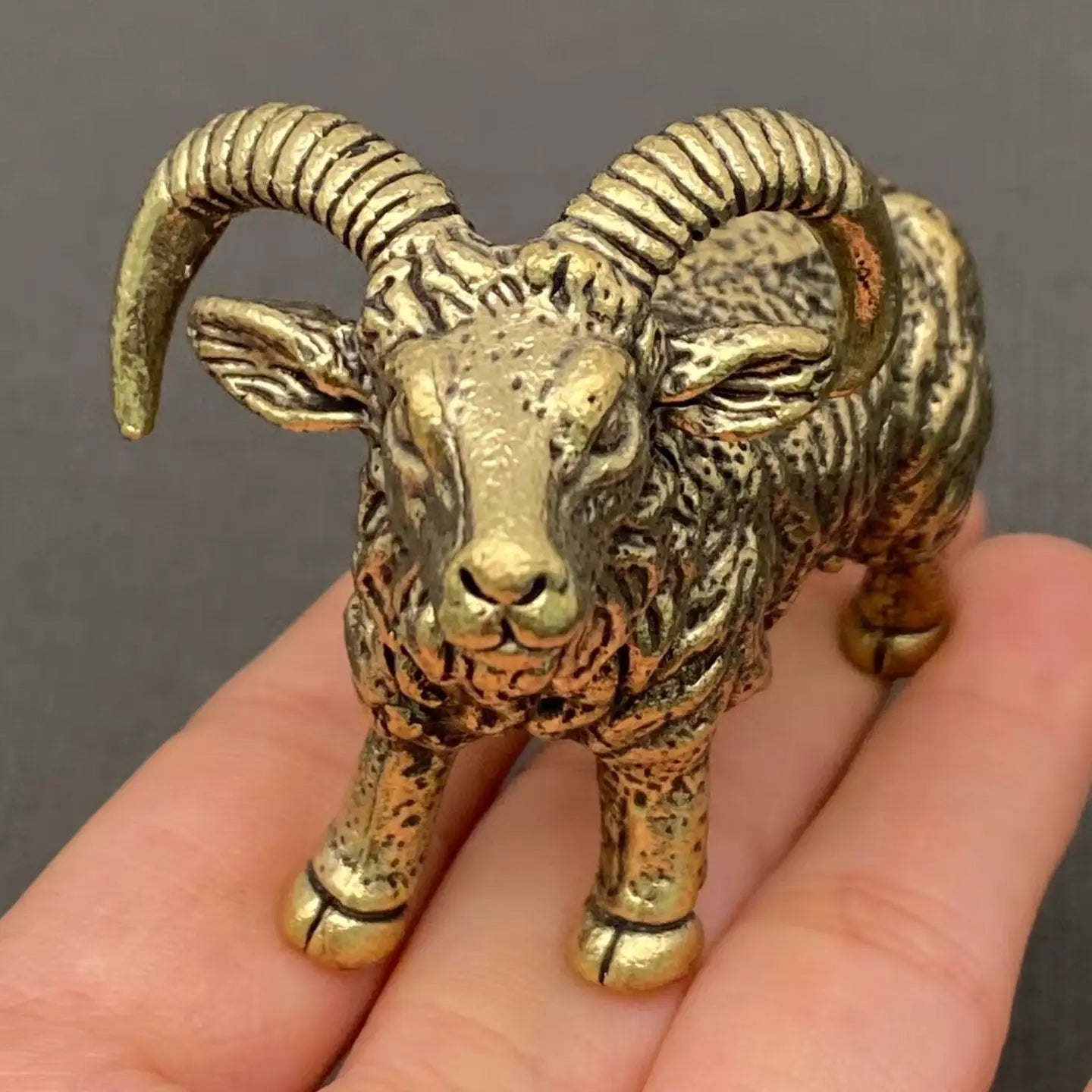 The Resilient Presence miniature brass ram with coiled horns standing firm, showing warm aged patina and textured detail on a wooden surface, from the WILLAGE Tiny Objects collection