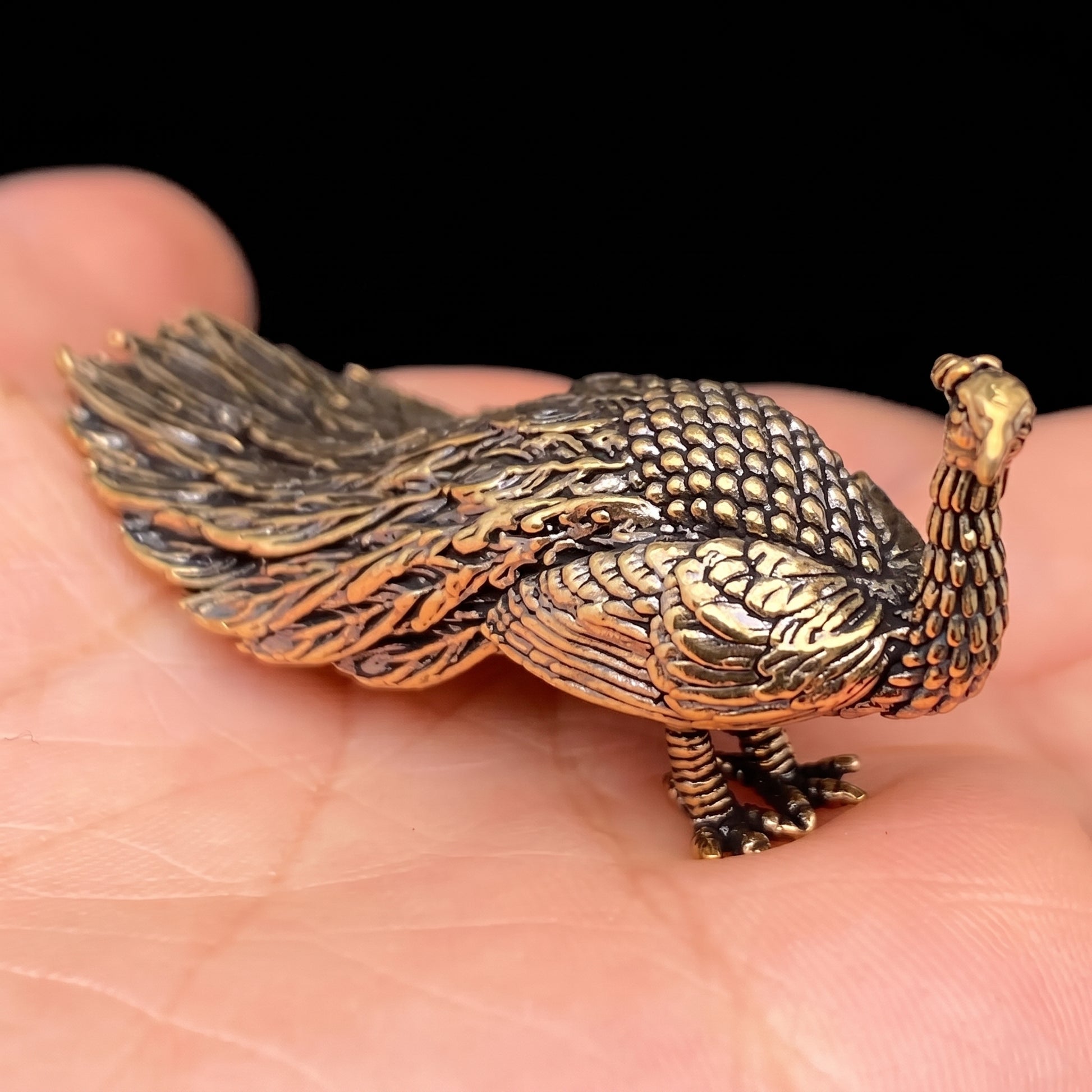 Small brass peacock figurine resting in an open palm, showing its sweeping tail feathers and detailed scaled neck