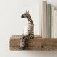 The Resting Zebra hand-carved wooden shelf sitter perched on a rustic wood shelf next to books, showing painted stripe detail and seated pose, carved by Kamba tribe artisans in Kenya