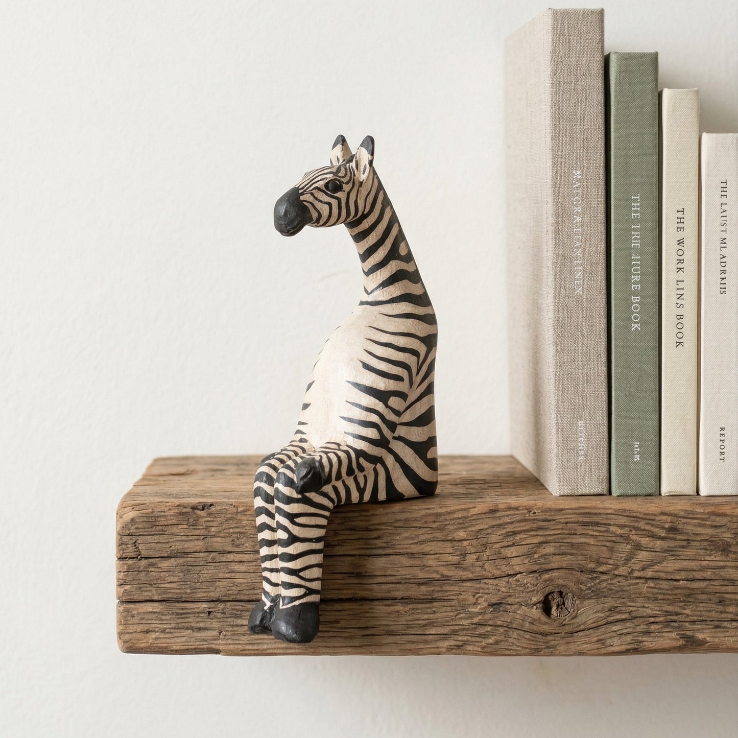 The Resting Zebra hand-carved wooden shelf sitter perched on a rustic wood shelf next to books, showing painted stripe detail and seated pose, carved by Kamba tribe artisans in Kenya