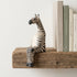 The Resting Zebra hand-carved wooden shelf sitter perched on a rustic wood shelf next to books, showing painted stripe detail and seated pose, carved by Kamba tribe artisans in Kenya