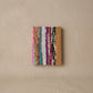 Pocket journal with a woven cover of multicolour upcycled sari strips in pink, teal, white, plum and green, with a tan leather edge, against a plaster wall