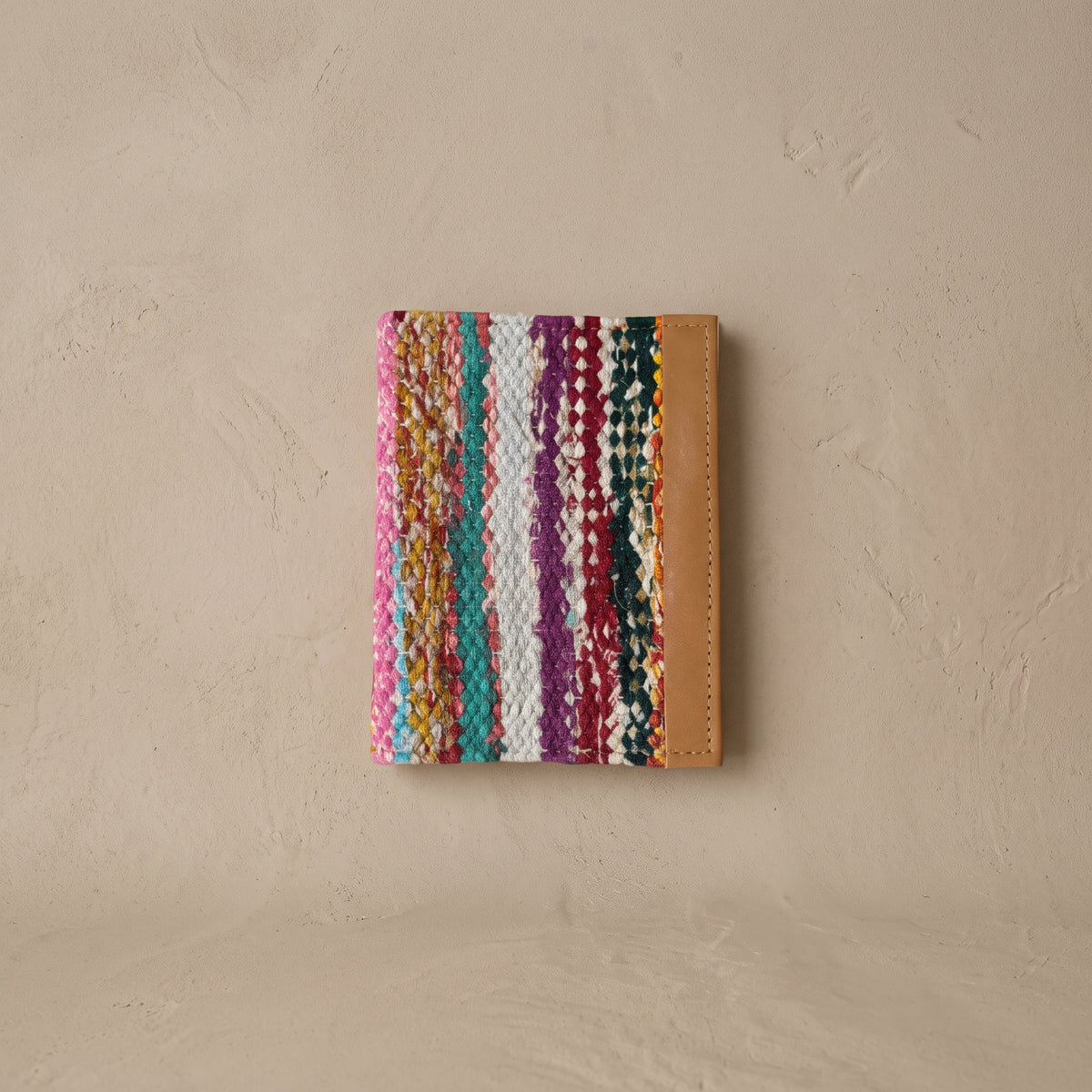 Pocket journal with a woven cover of multicolour upcycled sari strips in pink, teal, white, plum and green, with a tan leather edge, against a plaster wall