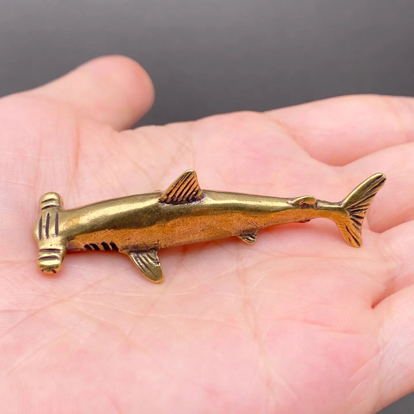The Silent Navigator miniature brass hammerhead shark held in hand to show scale, with streamlined body and golden patina