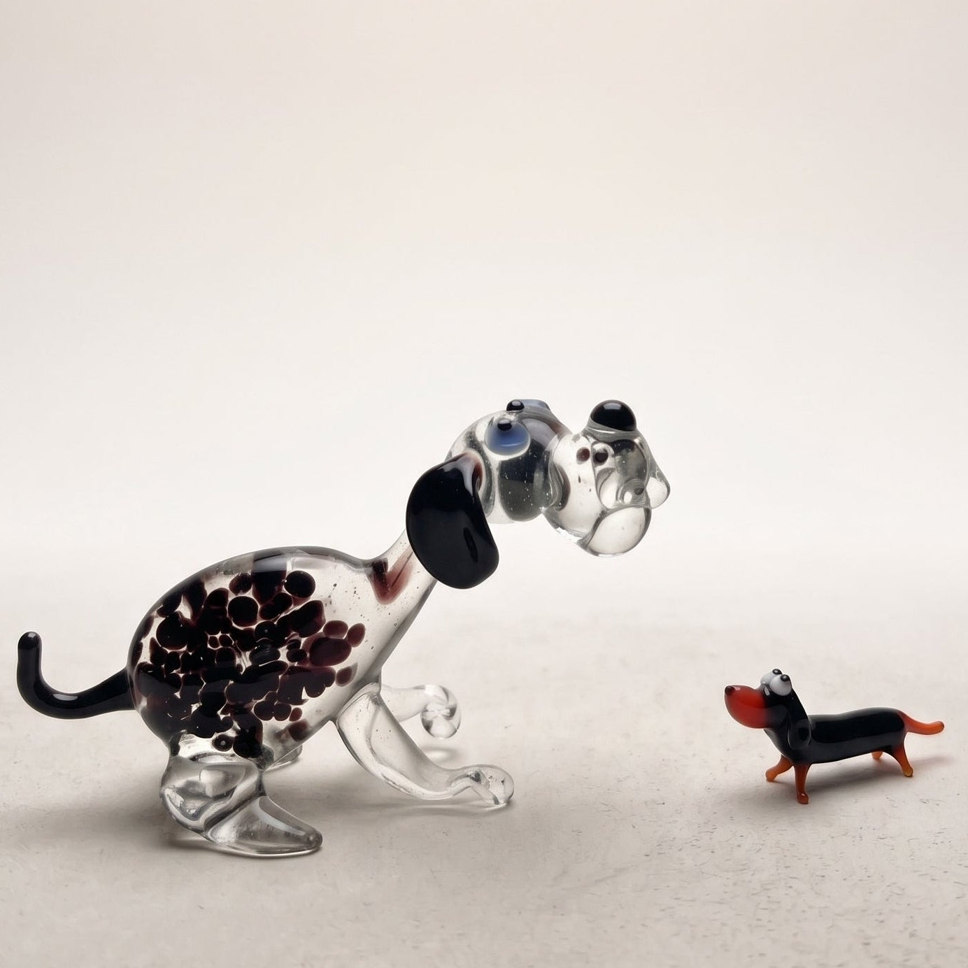 The Spotted Sprinter lampworked glass dog figurine with purple-black spots in clear glass alongside a miniature glass dachshund, handcrafted in L'viv Ukraine by Glass by Iness, from WILLAGE