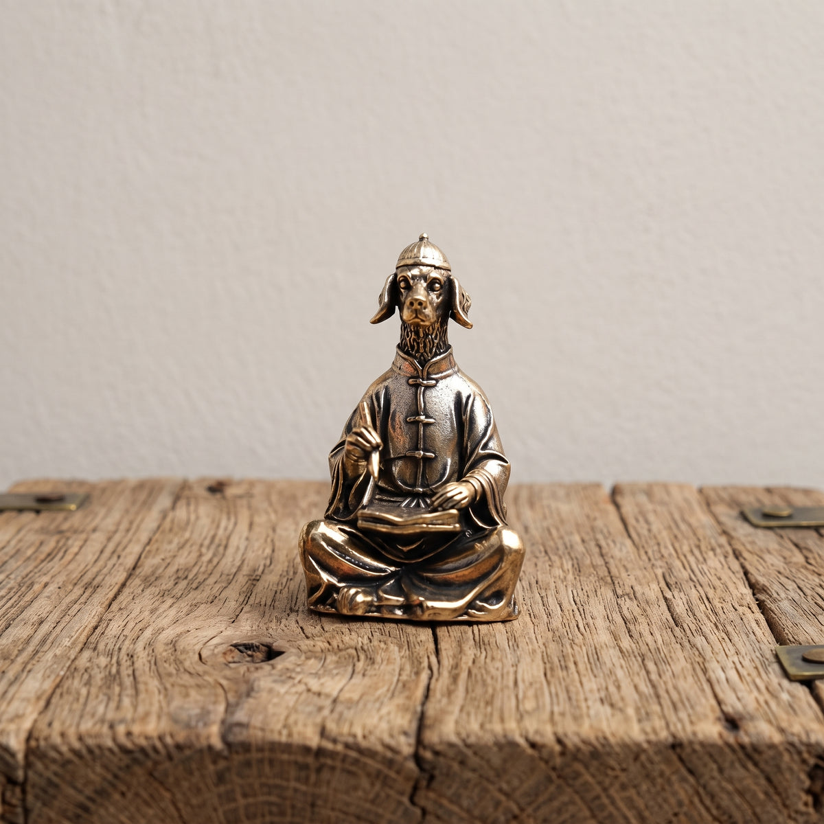Solid brass figurine of a dog in a scholar's robe and round cap, seated cross-legged with a brush raised over a book, on a weathered wood surface
