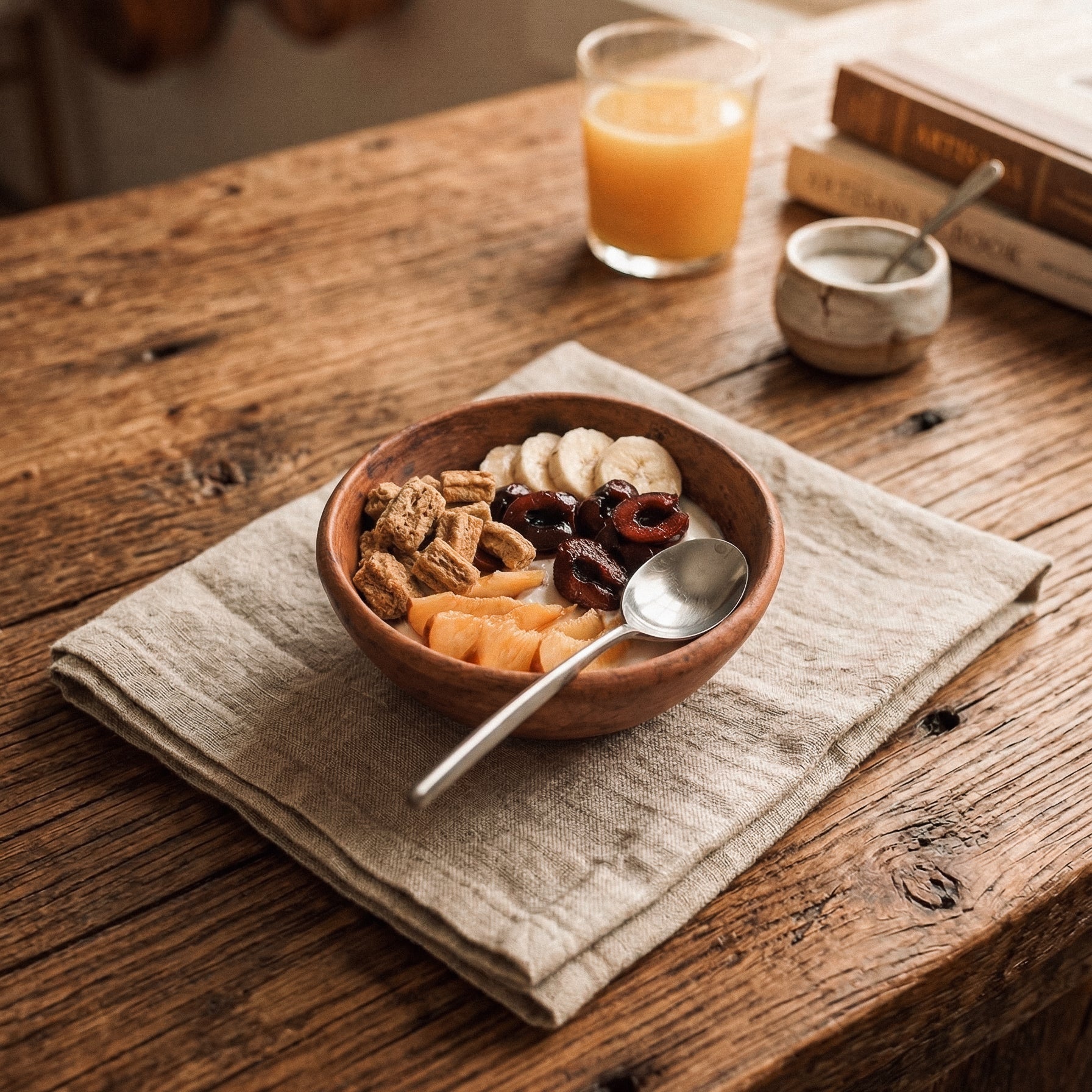 Sung cereal bowl in warm brown-toned black clay filled with cereal, banana, cherries, and fruit on a linen napkin and rustic wooden table with orange juice, handcrafted Lyrnai pottery from Meghalaya India, from WILLAGE