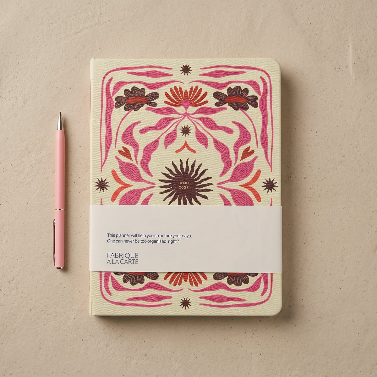 2027 weekly diary with a butter-yellow textured cover printed in magenta, rust and brown folk-art florals around a central dark sun, shown with its paper belly band