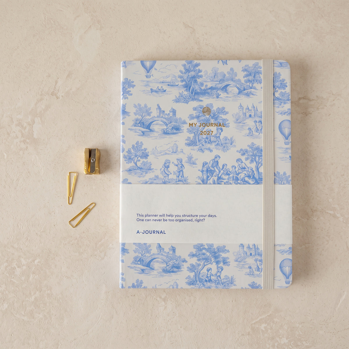 2027 planner with a Delft blue toile cover showing hot-air balloons, stone bridges and picnic scenes, gold-foil title, and a cream elastic band, on a pale plaster surface