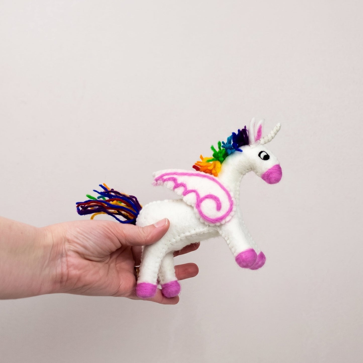 Rainbow-maned felted unicorn held in a hand to show its size and colourful wool tail