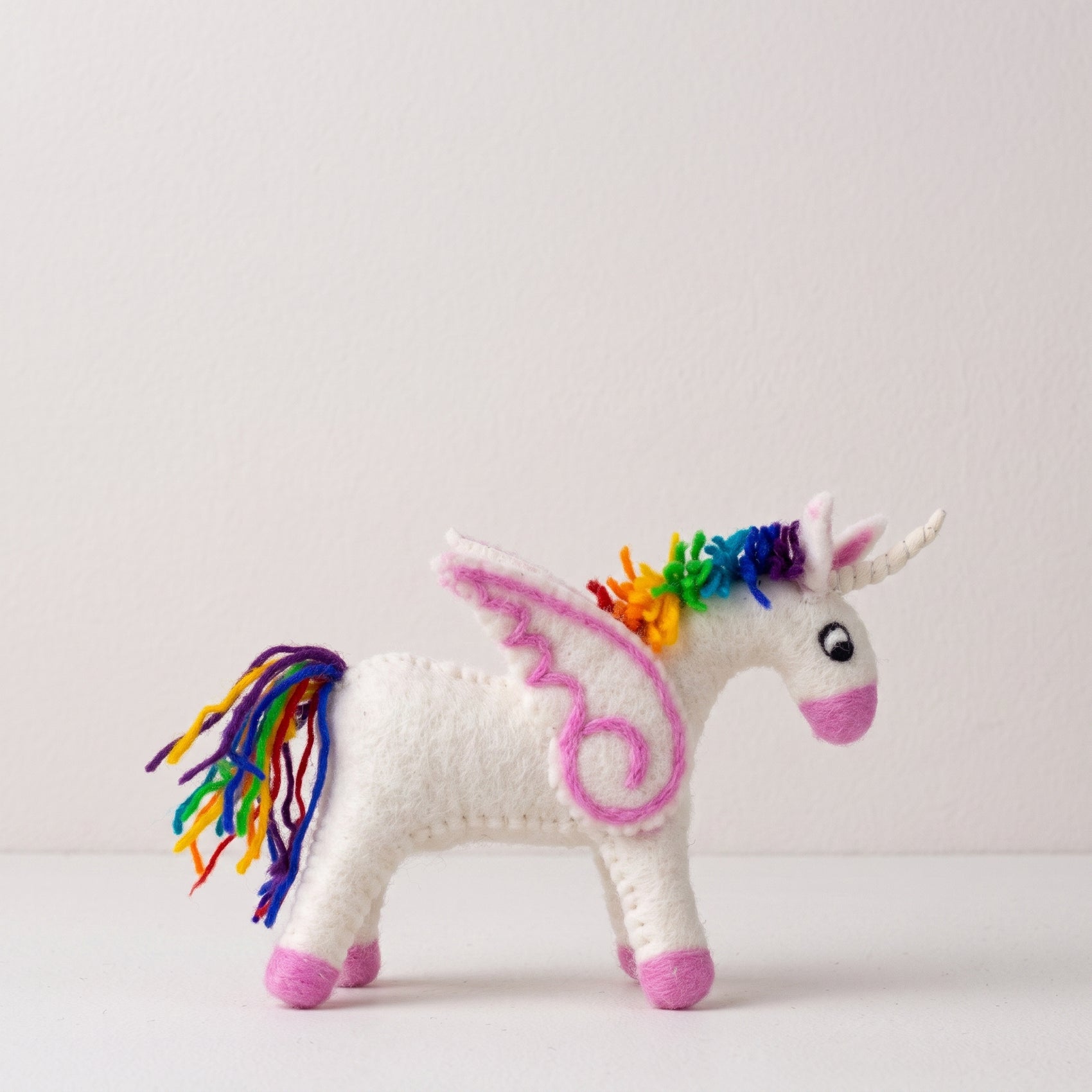 Hand-felted white unicorn with a rainbow mane and tail, pink muzzle and hooves, and felt wings, on a white background