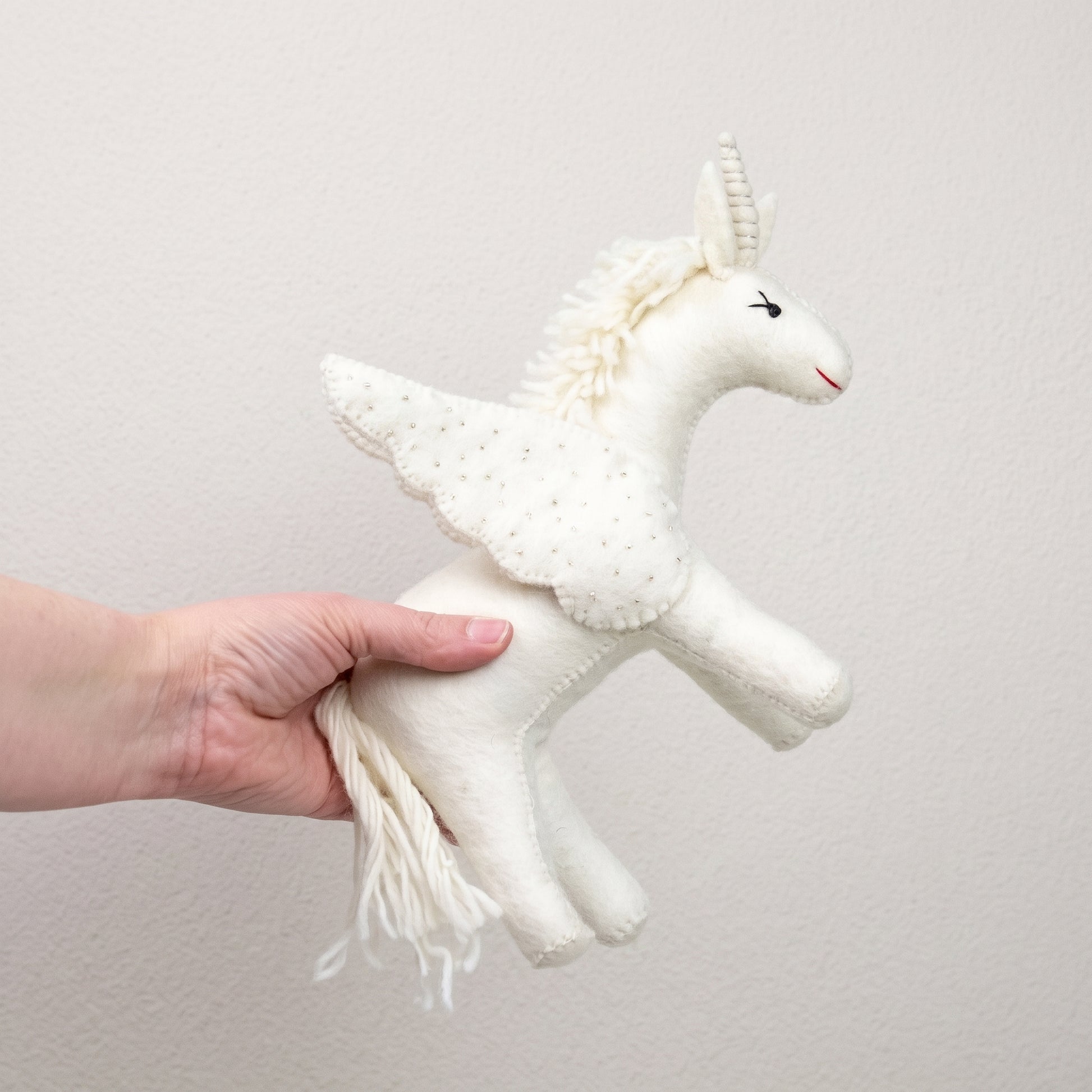 White felted unicorn held in a hand, showing its beaded wings and flowing wool tail