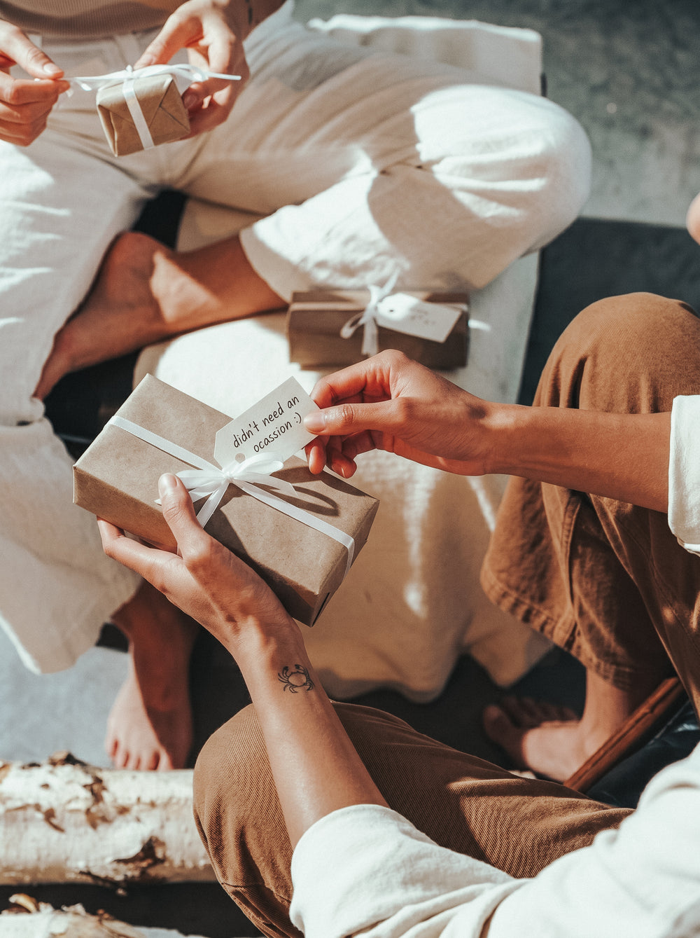Two people sitting together in warm sunlight, exchanging small gifts wrapped in kraft paper and tied with white ribbon, one tag reading 'didn't need an occasion :)'"