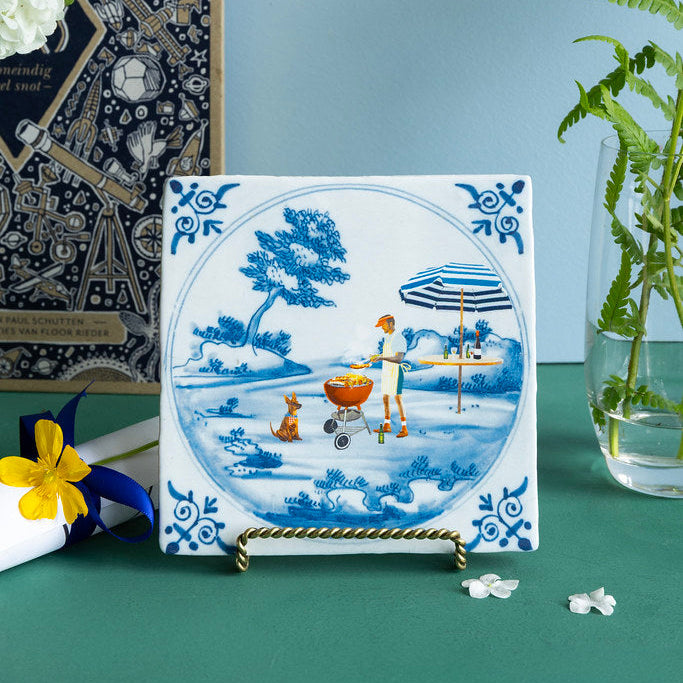 Decorative tile with a blue and white design of a man with a dog and a baby carriage, placed on a stand against a blue wall with plants.